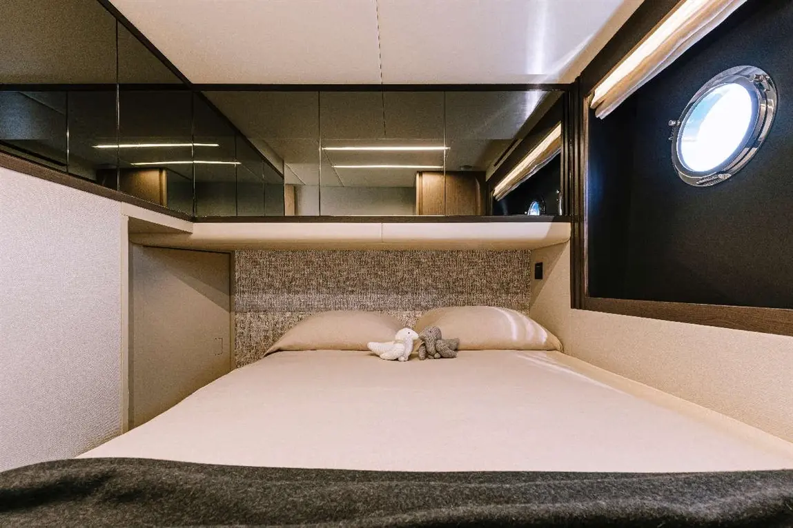 Cosy boat cabin with a double bed, plush pillows, and a decorative toy. Features a circular window and mirrored panels.